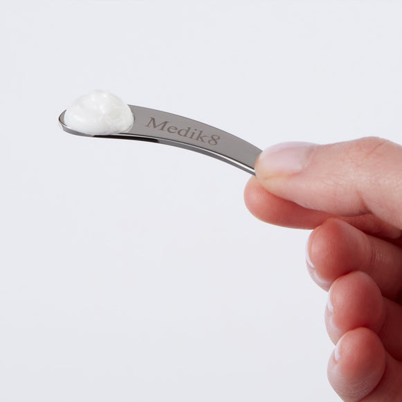 Une main tient une petite spatule incurvée en métal argenté, gravée du logo "Medik8". Une petite noisette de crème blanche est visible à l'extrémité de la spatule.-hover-47