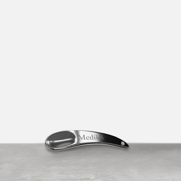 Une spatule en métal argenté repose sur une surface grise. La spatule courbée porte le logo "Medik8" gravé sur son manche.-47
