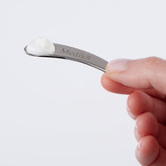 Une main tient une petite spatule incurvée en métal argenté, gravée du logo "Medik8". Une petite noisette de crème blanche est visible à l'extrémité de la spatule.