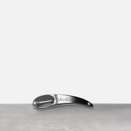 Une spatule en métal argenté repose sur une surface grise. La spatule courbée porte le logo "Medik8" gravé sur son manche.